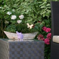 Rattan Beistelltisch mit Gartendekoration. Im Hintergrund Sträucher und Hortensien.