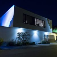 Nachtaufnahme - Neu gestalteter Vorgarten mit Bepflanzung, modernes Gebäude in blau beleuchtet.