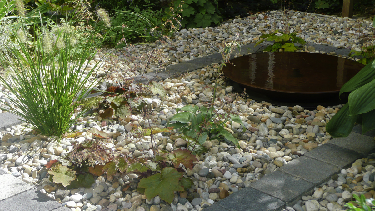 Im Zentrum des neu gestalteten Innenhofs Kiesbeet mit flachem Wasserspiel, bepflanzt mit Funkien, Heuchera und Ziergras, eingerahmt von einer Reihe Pflasterstein.