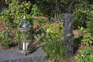 Neben einer Wasserstelle aus Naturstein im Beet, bepflanzt mit Gräsern und Schmuckblattpflanzen eine Gartenlampe aus Glas.