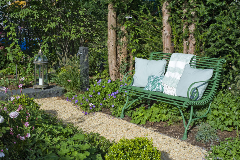 Gemütliches Gartendesign mit Sitzbank im bepflanzten Beet, mit Ziergras und blühenden Stauden. Daneben Wasserstelle aus Naturstein und Gartenlampion.
