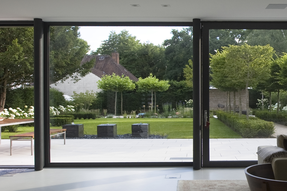 Blick durch die Glastür der Terrasse in den neu gestalteten Garten mit Quellsteinen, einer Allee und Hortensienbeeten.