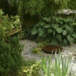 Flacher Stein als Wasserspiel im Kieselbett und kleine Holzbank umgeben von Funkien, blühenden Stauden und Rhododendron.