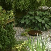 Flacher Stein als Wasserspiel im Kieselbett und kleine Holzbank umgeben von Funkien, blühenden Stauden und Rhododendron.