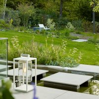 Planung und Gestaltung eines modernen Gartens in Hofheim - Wasserbecken in Betonoptik mit Trittstufen. Der Sonnenbereich des Gartens liegt hinter Beeten, bepflanzt mit Sträuchern und Stauden.