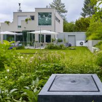 Planung und Gestaltung eines modernen Gartens in Hofheim - Blick aus dem bienenfreundlichen Bereich des Gartens, über die Rasenfläche zur Terrasse. Quellstein / Wasserspiel aus Basaltquader.