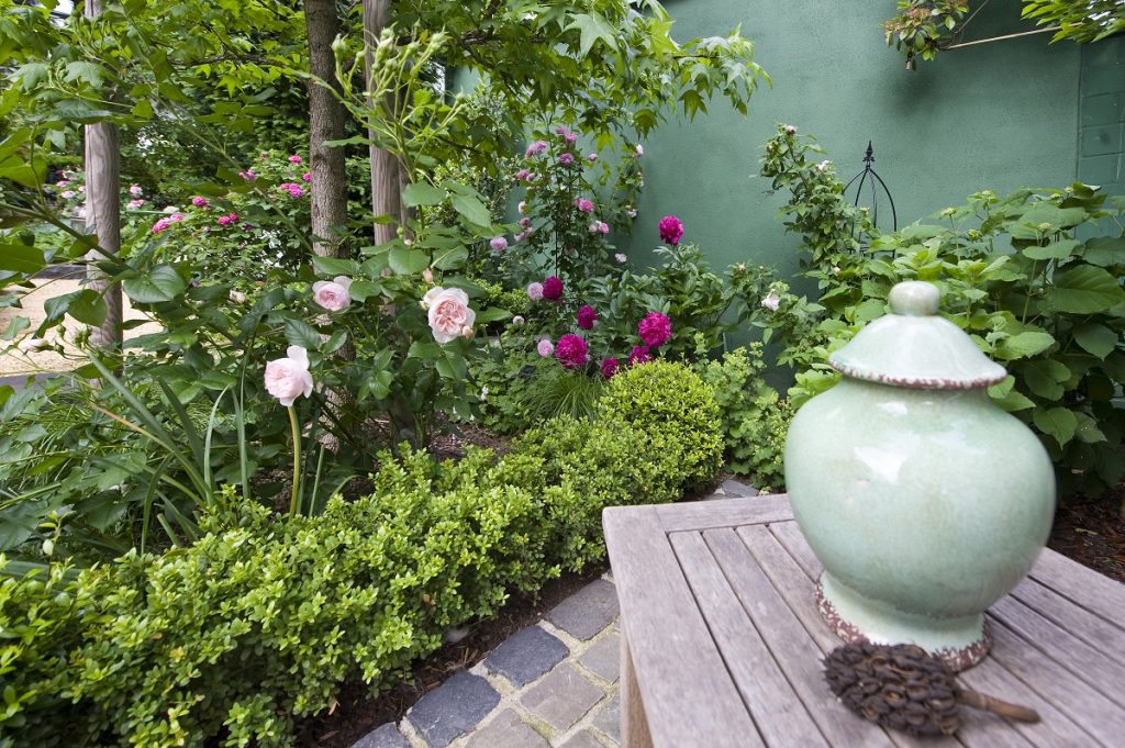 Vase als Gartendekoration auf Beistelltisch aus Holz, neben der Pflanzinsel mit weißer und zart rosa Rosen und Pfingstrosen.