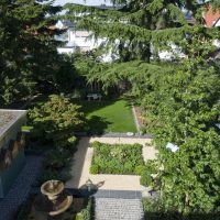 Blick von oben auf das neue Gartendesign. Gartengestaltung mit klassischem Springbrunnen aus Stein, einer Pflanzinsel mit Buchsaum und Stauden, umgeben von Kiesweg und Rasen unter hohen alten Bäumen.
