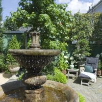 Klassischer Springbrunnen aus Stein, im Hintergrund gemütliche Liege neben Hortensien. Klassischer Garten mit sonnigen und schattigen Bereichen.