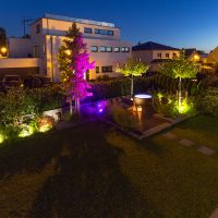 Nachtansicht eines modernen Gartens, Lounge Ecke mit Holzdielen, Beleuchtung der Bäume und Randbepflanzung, Trittplatten in der Rasenfläche