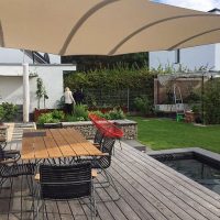 Möblierte Terrasse mit Holzboden, darüber ein Sonnensegel. Abtretung zur Rasenfläche durch Gabionen Gartenbank und Wasserbecken.