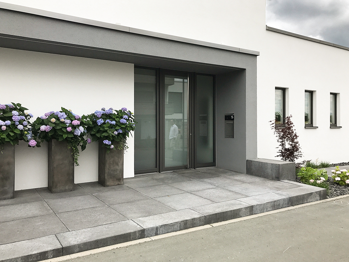 Moderne Architektur und minimaloistischer Vorgarten im Eingangsbereich. Große Bodenplatten in grau, ebenso in Betonoptik Pflanzkübel mit Hortensien.