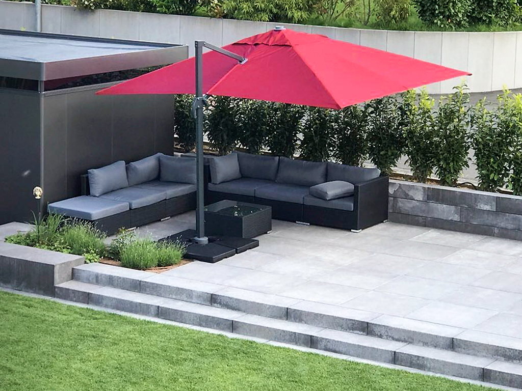 Großer, roter Sonnenschirm über hochwertige Gartenmöbel auf erhöhter Sitzfläche aus grauem Stein. Pflanzinsel mit Lavendel und Ziergras, sowie Gartendekoration